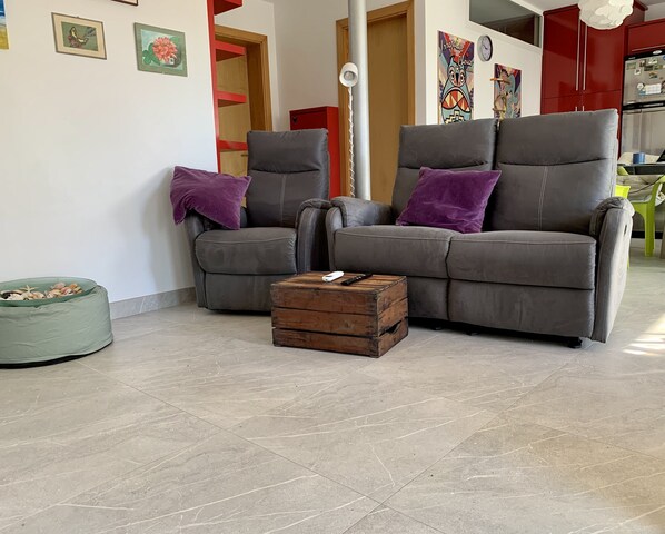 Een woonkamer met twee grijze banken, een houten kist en een groene relaxfauteuil.