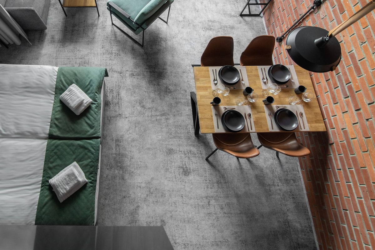 A hotel room with a bed, two green pillows, a wooden table set for dining, and a brick wall.