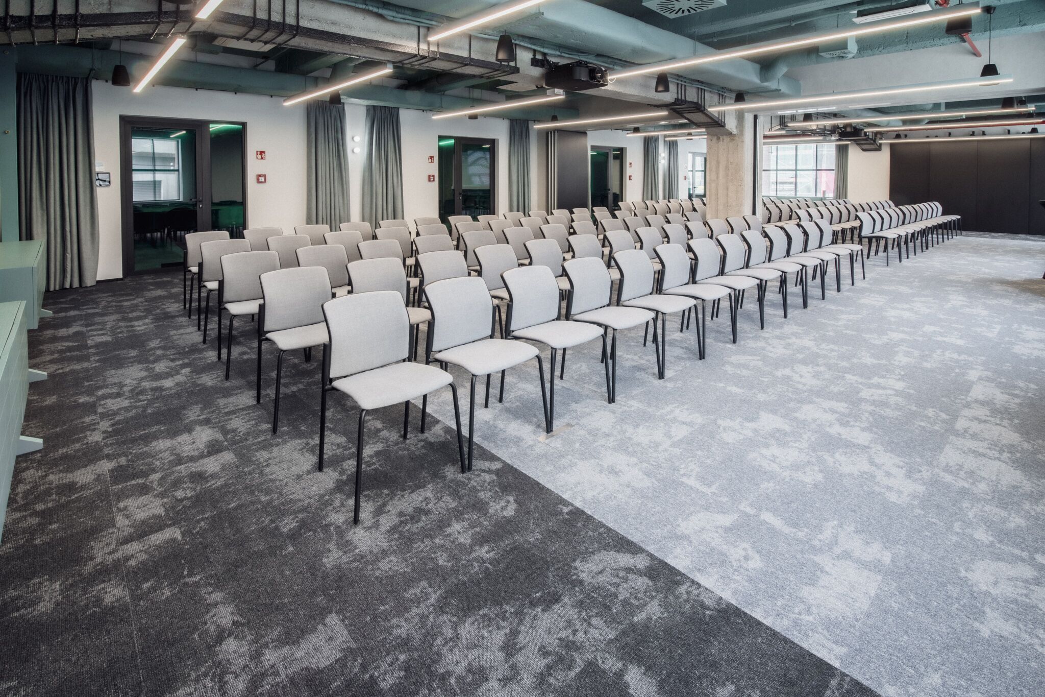 Ett konferensrum med rader av stolar, ett heltäckningsmatta och ett modernt tak med synliga rör och belysning.