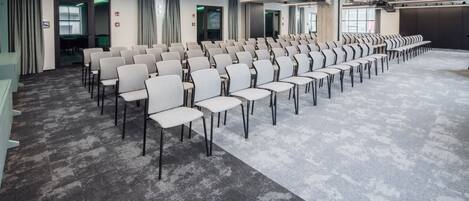A conference room with rows of chairs, a carpeted floor, and a modern ceiling with exposed pipes and lighting.