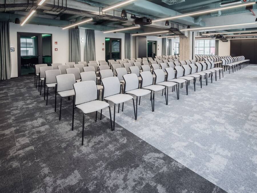 Une salle de conférence avec des rangées de chaises, un sol recouvert de moquette et un plafond moderne avec des tuyaux apparents et un système d’éclairage contemporain.