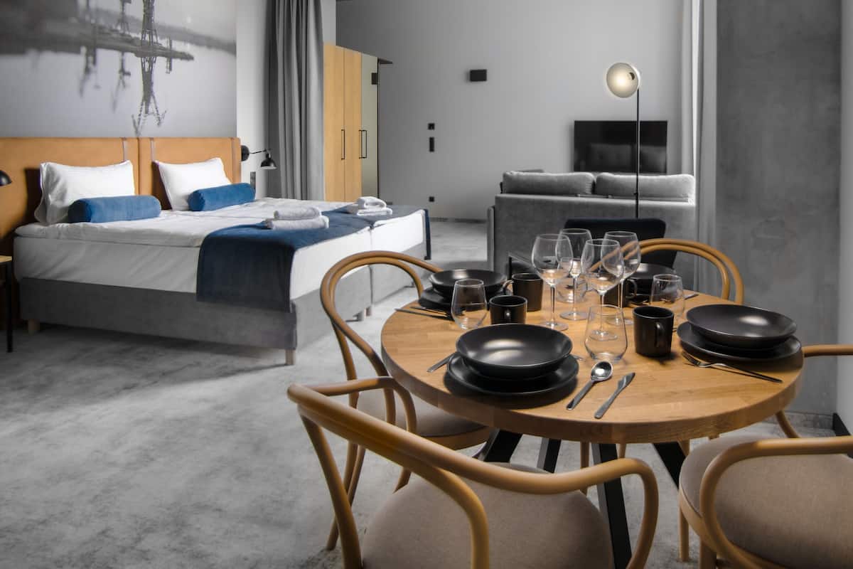 A modern hotel room with a neatly made bed, a dining table set for four, and a sofa in the background.