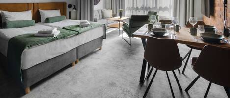 Una habitación de hotel moderna con una cama bien hecha, una zona de comedor con sillas y una mesa preparada para dos personas, y un gran ventanal con cortinas.
