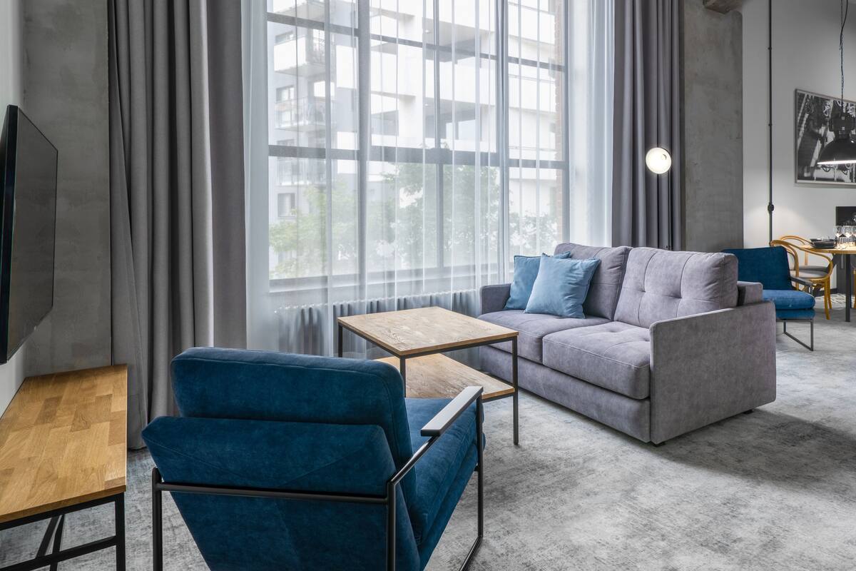 A modern living room with a grey sofa, blue armchairs, a wooden coffee table, and a large window with sheer curtains.
