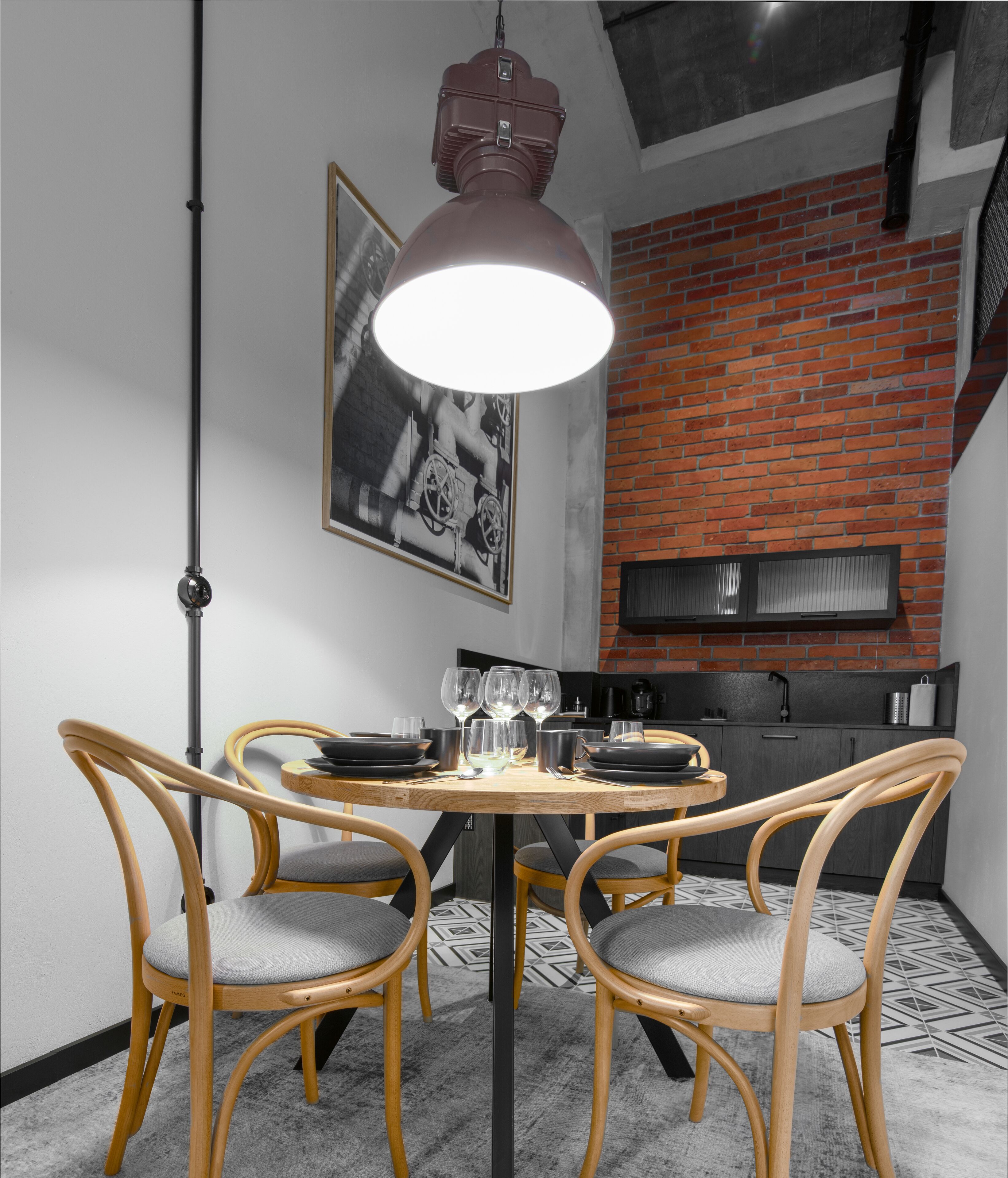 A dining area with a round table, four wooden chairs, and a pendant light. A brick wall and a kitchen with a black range hood are visible in the background.