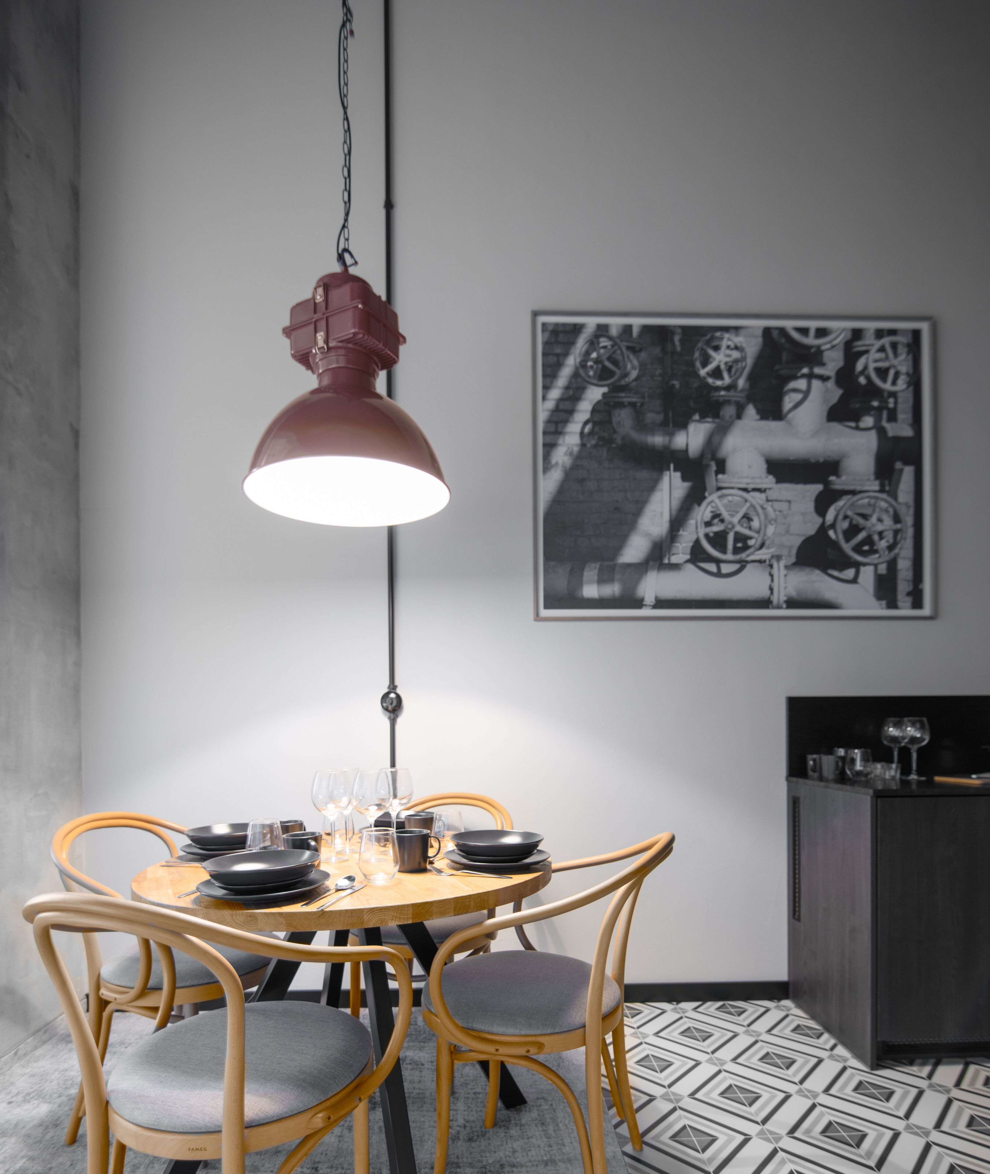 Un coin repas avec une table ronde dressée pour quatre personnes, entourée de chaises en bois avec des coussins gris. Un luminaire suspendu est placé au-dessus de la table, et une photographie en noir et blanc est accrochée au mur.