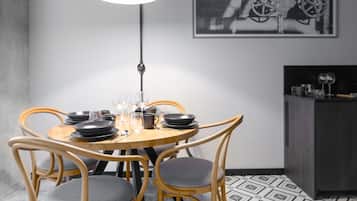 A dining area with a round table set for four, surrounded by wooden chairs with grey cushions. A pendant light hangs above the table, and a black and white photograph is displayed on the wall.