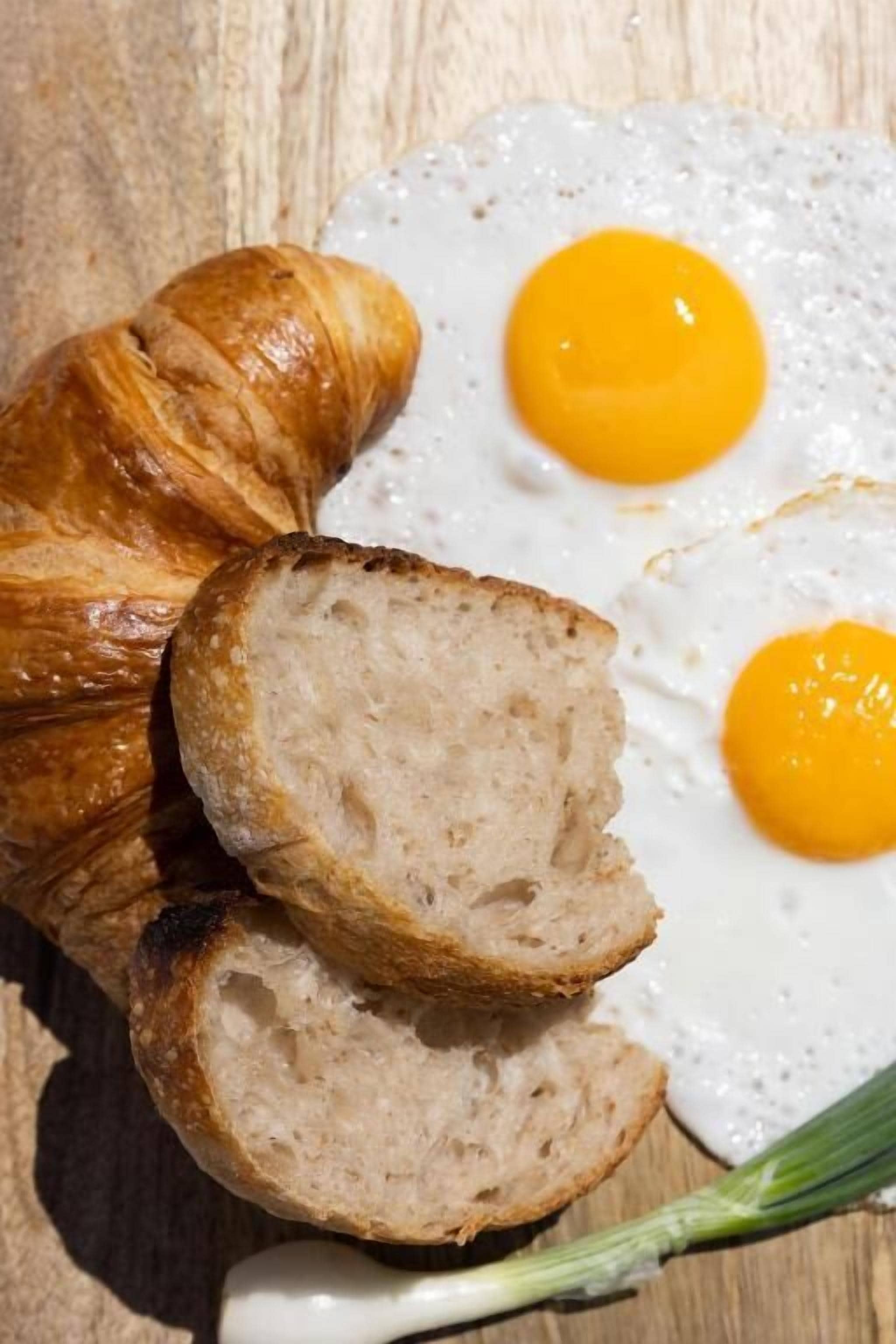 A croissant, two slices of bread, and two fried eggs on a wooden surface.