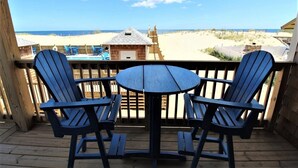 Una terrazza in legno con due poltrone Adirondack e un tavolino, affacciata su una spiaggia con dune sabbiose e una struttura in legno all'orizzonte.