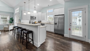 A spacious kitchen with white cabinets, a central island with stools, stainless steel appliances, and pendant lighting. - Home on the Banks at Beacon Villas 4 Bedroom Townhouse (Corolla)
