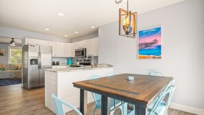 A modern kitchen with white cabinets, stainless steel appliances, and a wooden dining table with blue chairs. - Amina's Cottage 3 BR in KDH + Pets Stay Free (Kill Devil Hills)