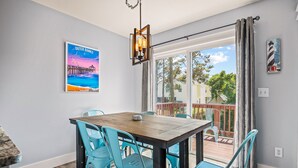 A dining area with a wooden table and turquoise chairs, a pendant light, and a view of the outdoors through large windows. - Amina's Cottage 3 BR in KDH + Pets Stay Free (Kill Devil Hills)