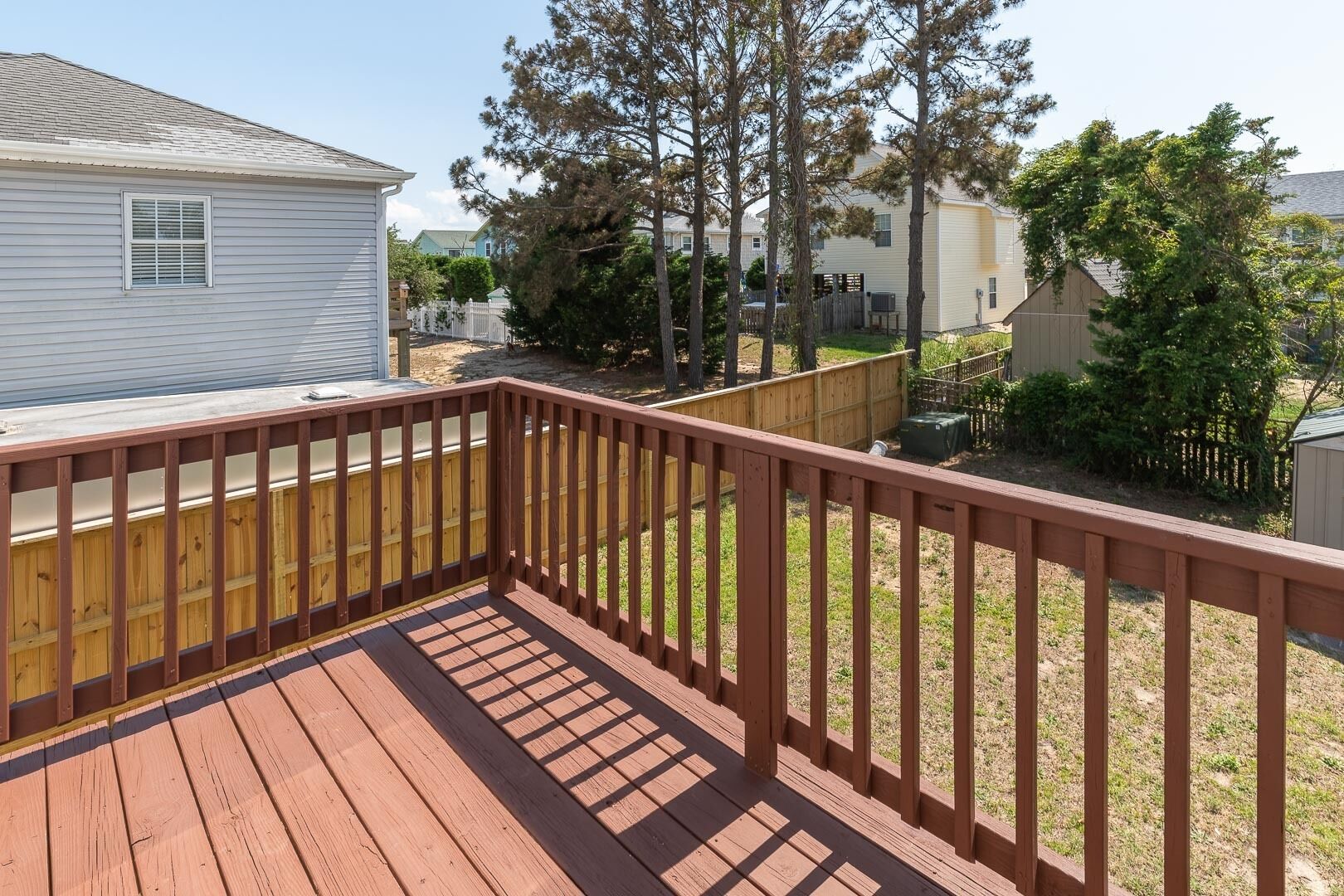 Une terrasse en bois avec une rambarde, entourée d’une cour clôturée, agrémentée d’arbres et avec un bâtiment visible au loin.
