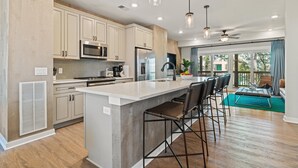 A modern kitchen with a large island, bar stools, and a dining area with a view. - Sea Therapy at Cambridge Cove (Kill Devil Hills)
