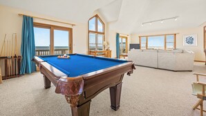 Une table de billard dans une pièce donnant sur la plage, avec un canapé et une fenêtre équipée de stores.