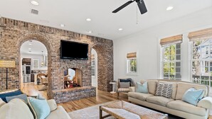 A spacious living room with a brick fireplace, a large flat-screen TV mounted above it, and a comfortable seating area with a sofa and a bench. - Family Home | Beach Access | Private Guest House (Inlet Beach)