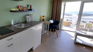 A modern kitchen with a sink, countertop, and cabinets. There is a dining area with a chair and a table. The room has a large window with a view of buildings and a balcony. - Beachlodge by the Sea | Honeymoon suite on the beach (Wijk aan Zee)