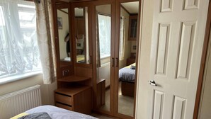 A bedroom with a bed, a wardrobe, a mirror, and a window with curtains. - Cozy 2 Bedroom Coastal Chalet in Hunstanton (King's Lynn)