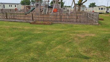 Un parque infantil con estructuras de madera y un área vallada, rodeado de césped y árboles.