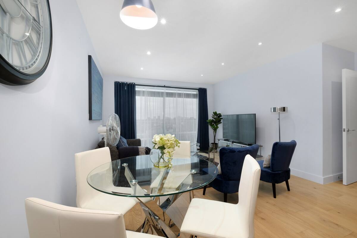 A modern living room with a glass dining table, white and blue chairs, a large window with curtains, a flat-screen TV, and a ceiling fan.