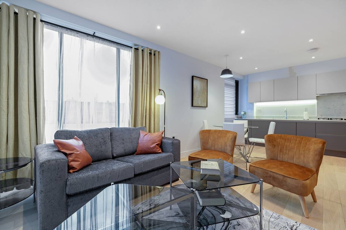 A modern living room with a grey sofa and two armchairs, a glass coffee table, and a view of the kitchen in the background.
