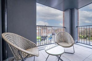 A balcony with two wicker chairs and a small table, overlooking a street with buildings and a clear sky.