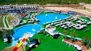 Una gran piscina con tobogán, rodeada de césped verde y sombrillas.