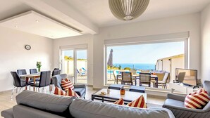 Amplia sala de estar con un sofá grande, una mesa de comedor y sillas. Hay un balcón con vista al mar.