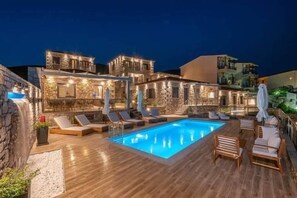 A poolside area with lounge chairs and a swimming pool, surrounded by stone buildings with warm lighting. - Lithoessa Luxury Apartments (Lemnos)