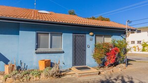 Een gelijkvloers huis met een blauwe gevel, een pannendak en een kleine voortuin met planten en een boom met rode bladeren.