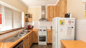 Een keuken met houten kastjes, een fornuis, een spoelbak en een koelkast. Op de koelkast staan bordjes met informatie over de warmwater voorziening en een rookverbod.