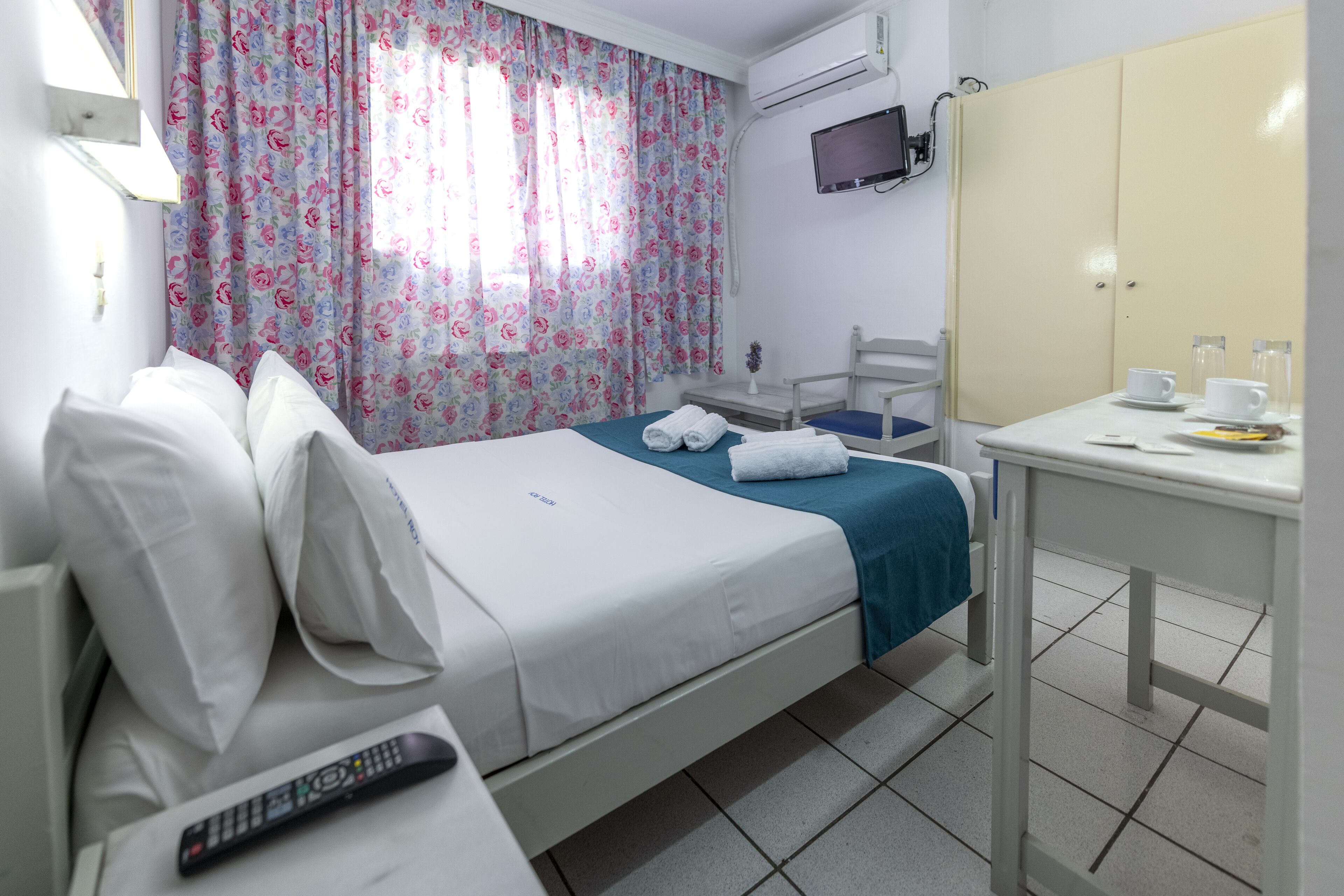 A hotel room with a bed, a table with a cup and a plate, a TV mounted on the wall, and a window with floral curtains.
