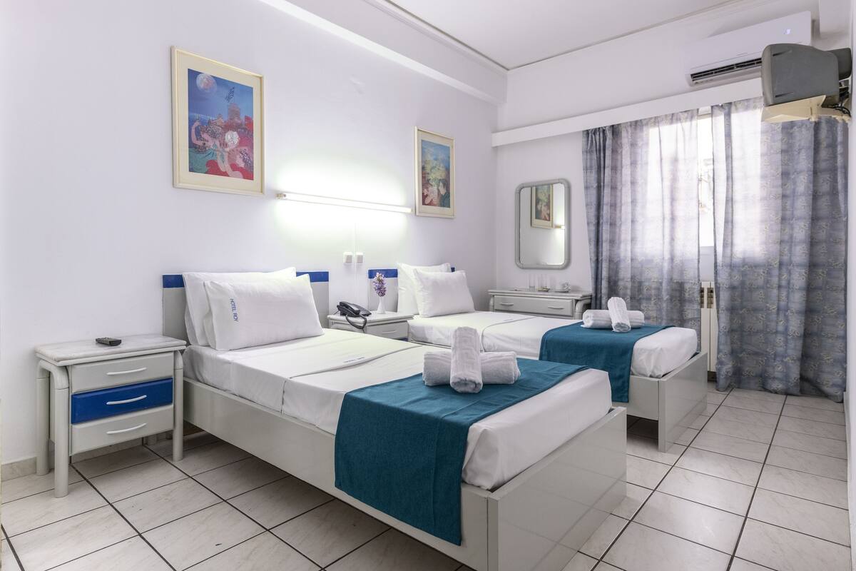 A hotel room with two single beds, a bedside table with a lamp, a small desk, and a window with curtains.