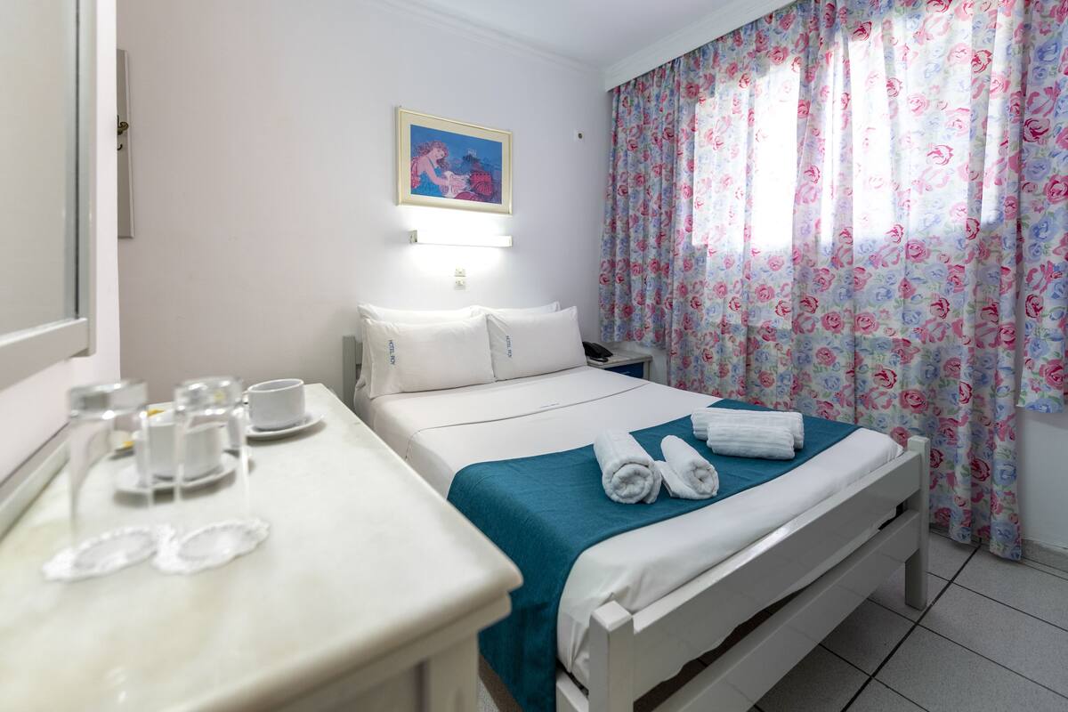 A hotel room with a bed, a bedside table with cups and plates, a window with floral curtains, and a painting on the wall.