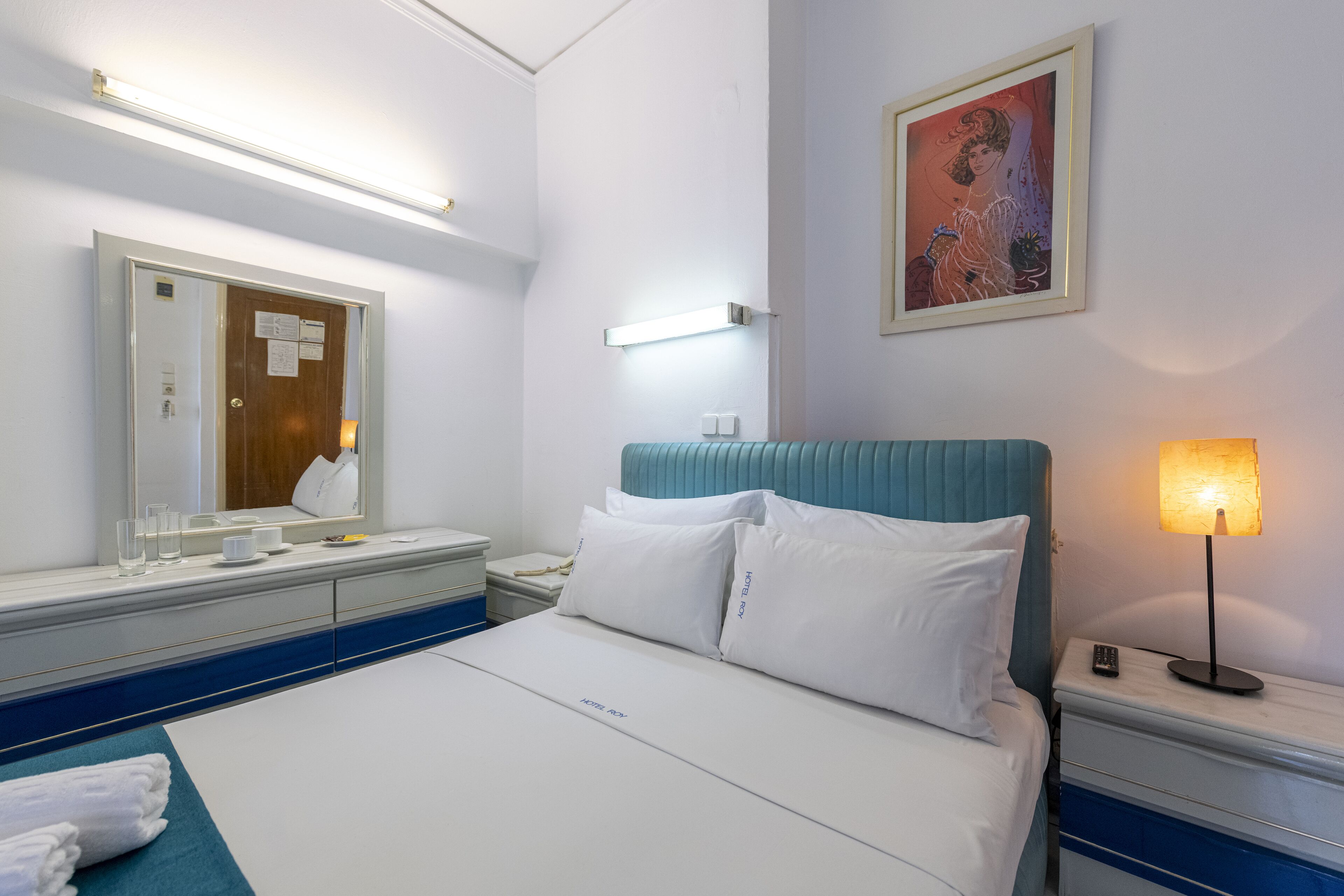 Hotellihuone, jossa on sänky, yöpöytä, sängyn vieressä oleva pöytä, peili, taulu ja lamppu.