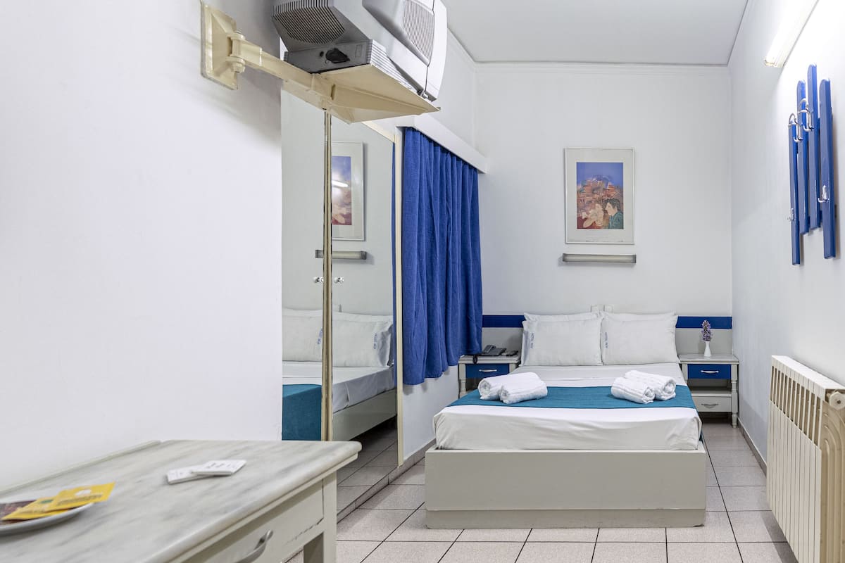 A hotel room with a bed, a desk, a chair, a mirror, a window with curtains, and a radiator.