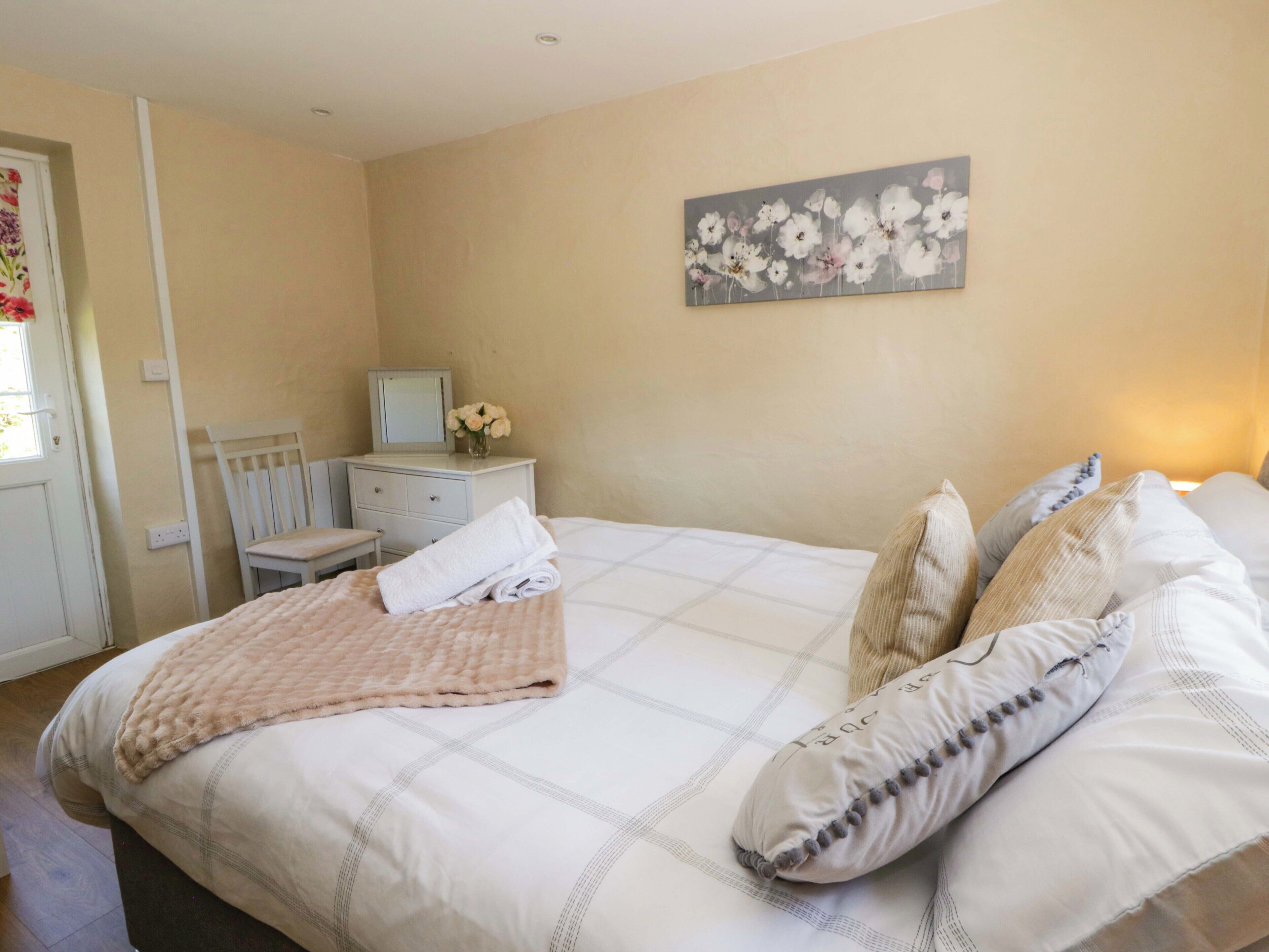 Une chambre à coucher avec un lit, des oreillers, une table de chevet, un miroir, une commode et un tableau représentant des fleurs accroché au mur.
