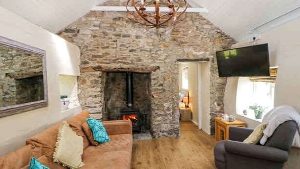 A cozy living room with a stone fireplace, a sofa with decorative pillows, a grey armchair, and a flat-screen TV mounted on the wall. - Cobwebs (Kilgetty)