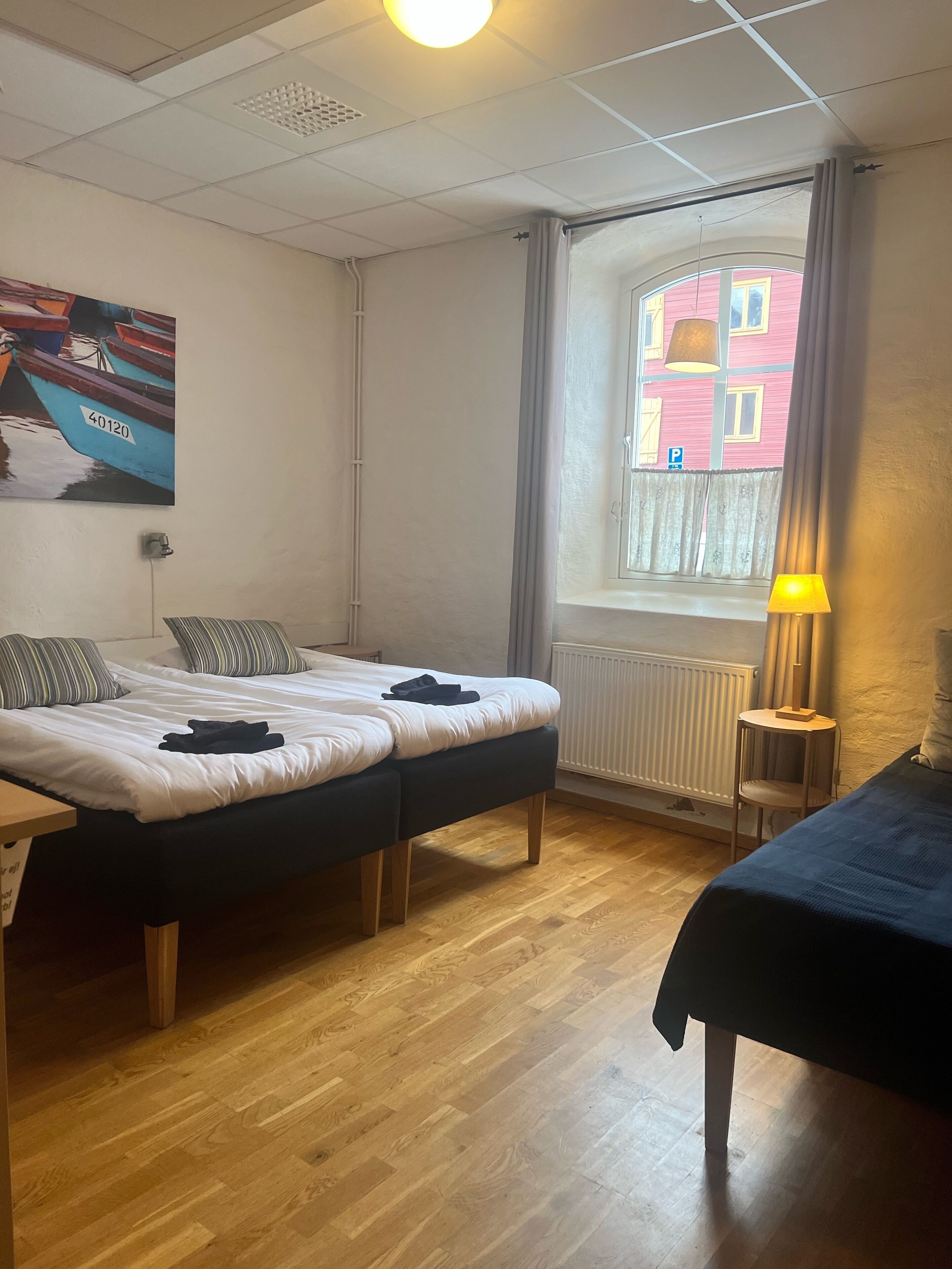 A room with two beds, a wooden floor, a window with a view of a pink building, and a painting of boats.