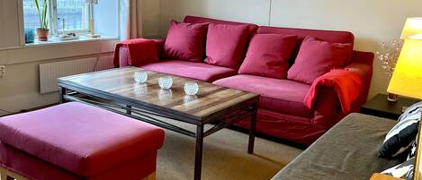 A living room with a red sofa, a wooden coffee table, and a grey ottoman. There is a window with a view of buildings outside.