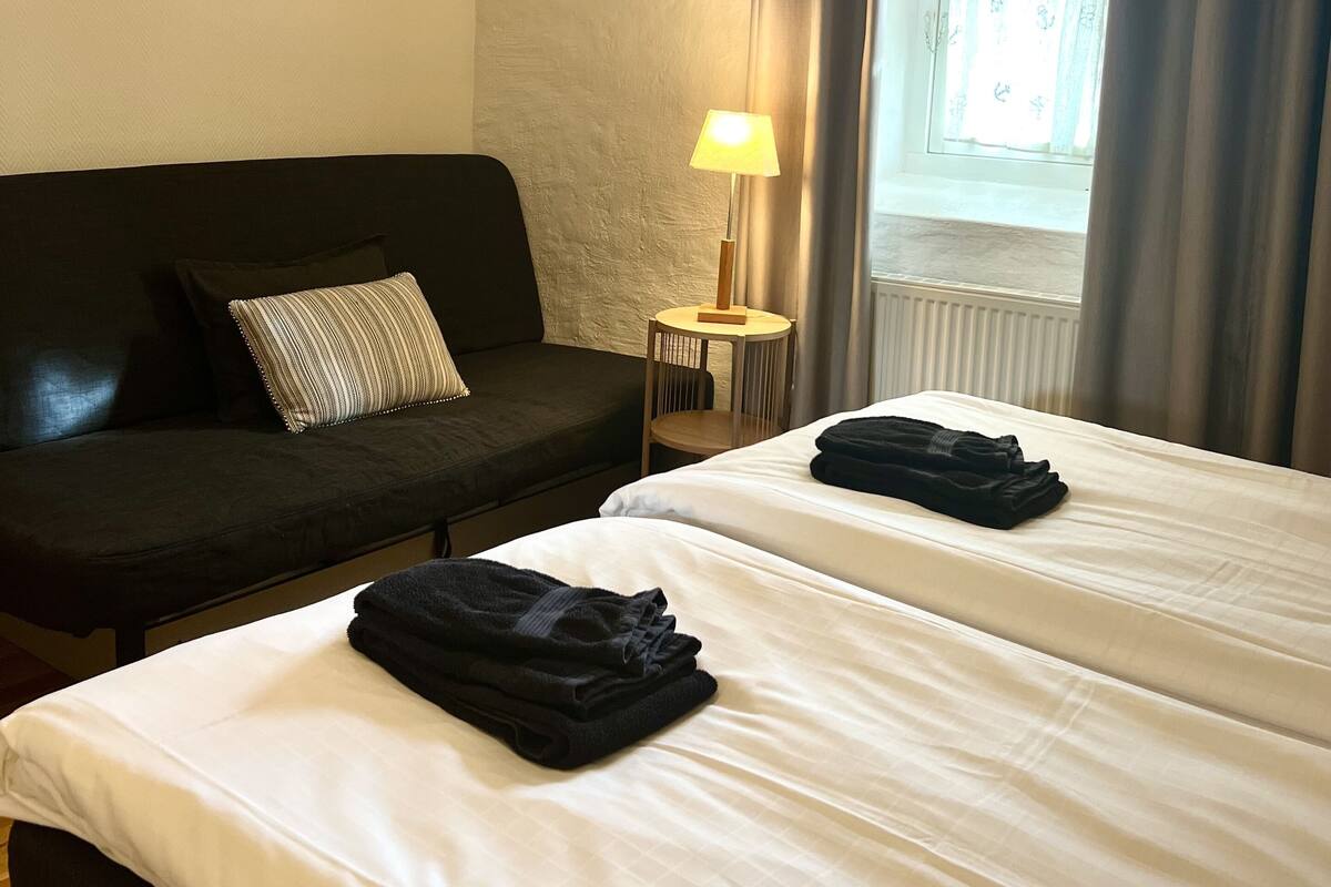 A hotel room with two beds, a dark-colored sofa, a small table with a lamp, and a window with curtains.