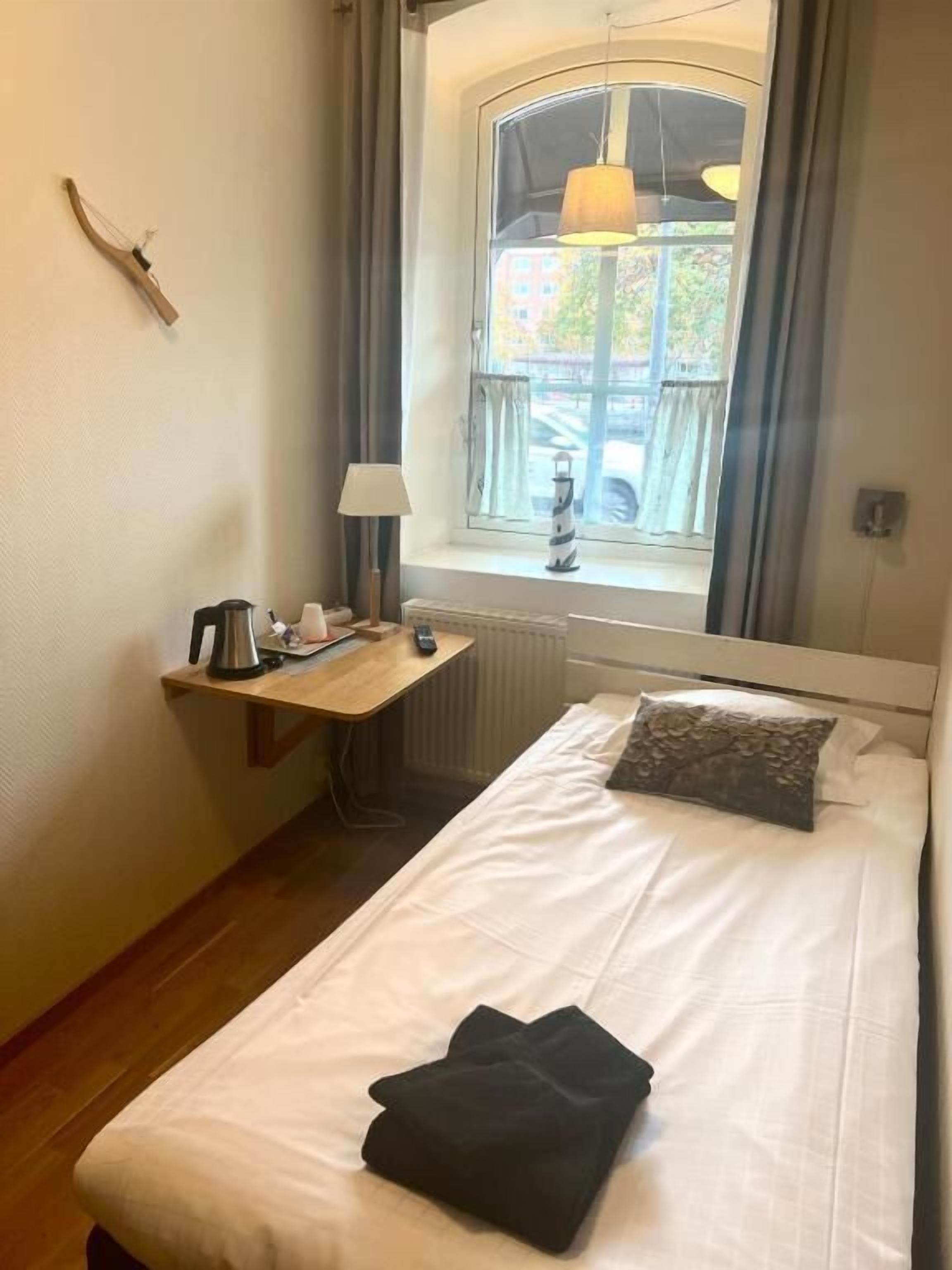 Un dormitorio pequeño y ordenado con una cama individual, una mesita de noche con lámpara, un radiador y una ventana con cortinas.