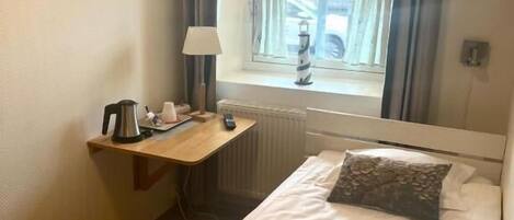 Un dormitorio pequeño y ordenado con una cama individual, una mesita de noche con lámpara, un radiador y una ventana con cortinas.