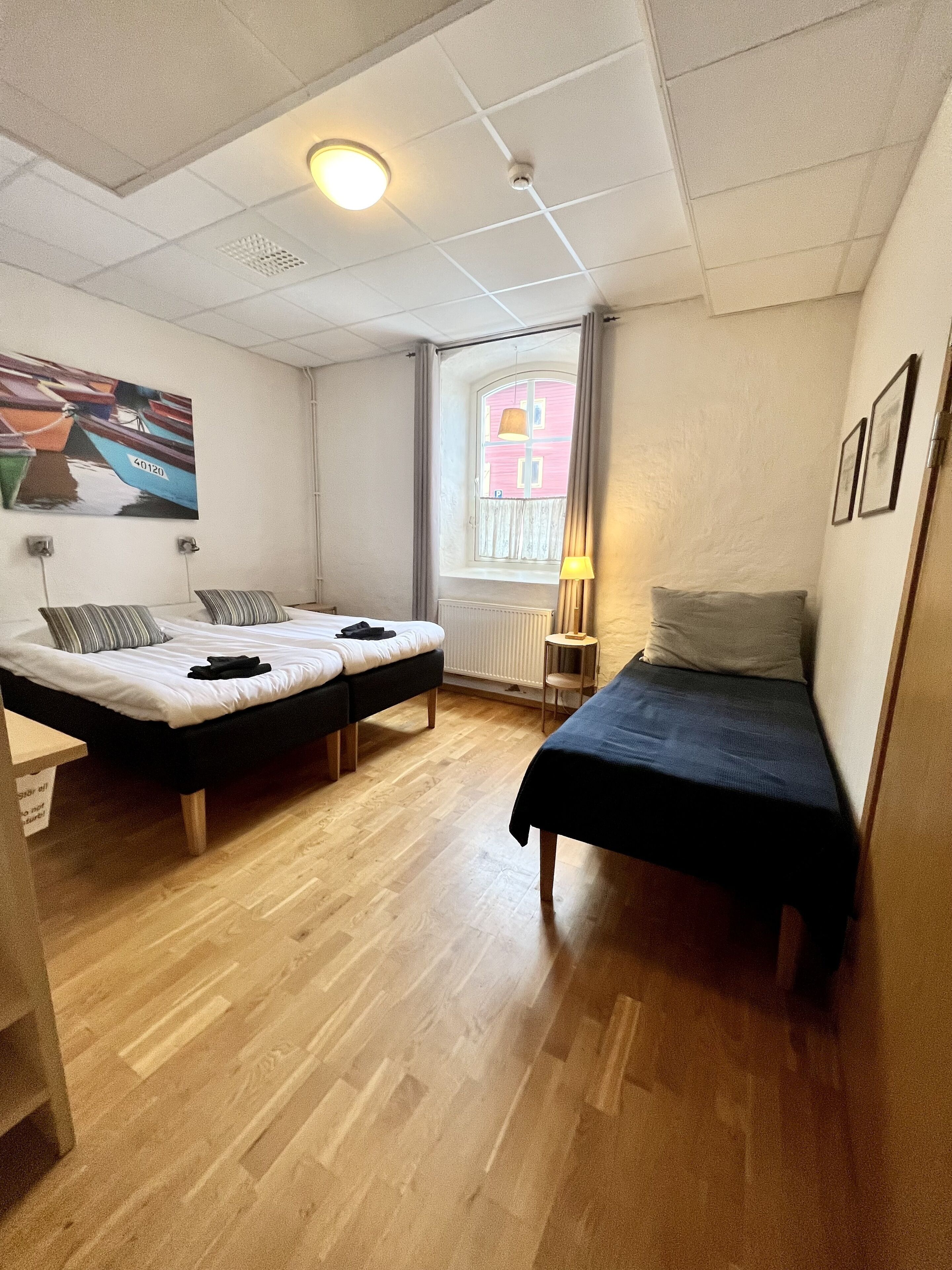A room with two beds, a window with a view of a pink building, a painting on the wall, and a lamp.