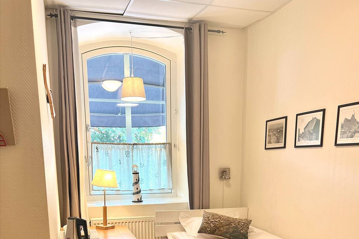 A small, well-lit room with a bed, a chair, a table with a lamp, and a window with curtains.
