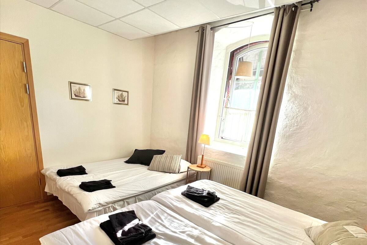 A hotel room with two beds, wooden flooring, a window with curtains, and a lamp.