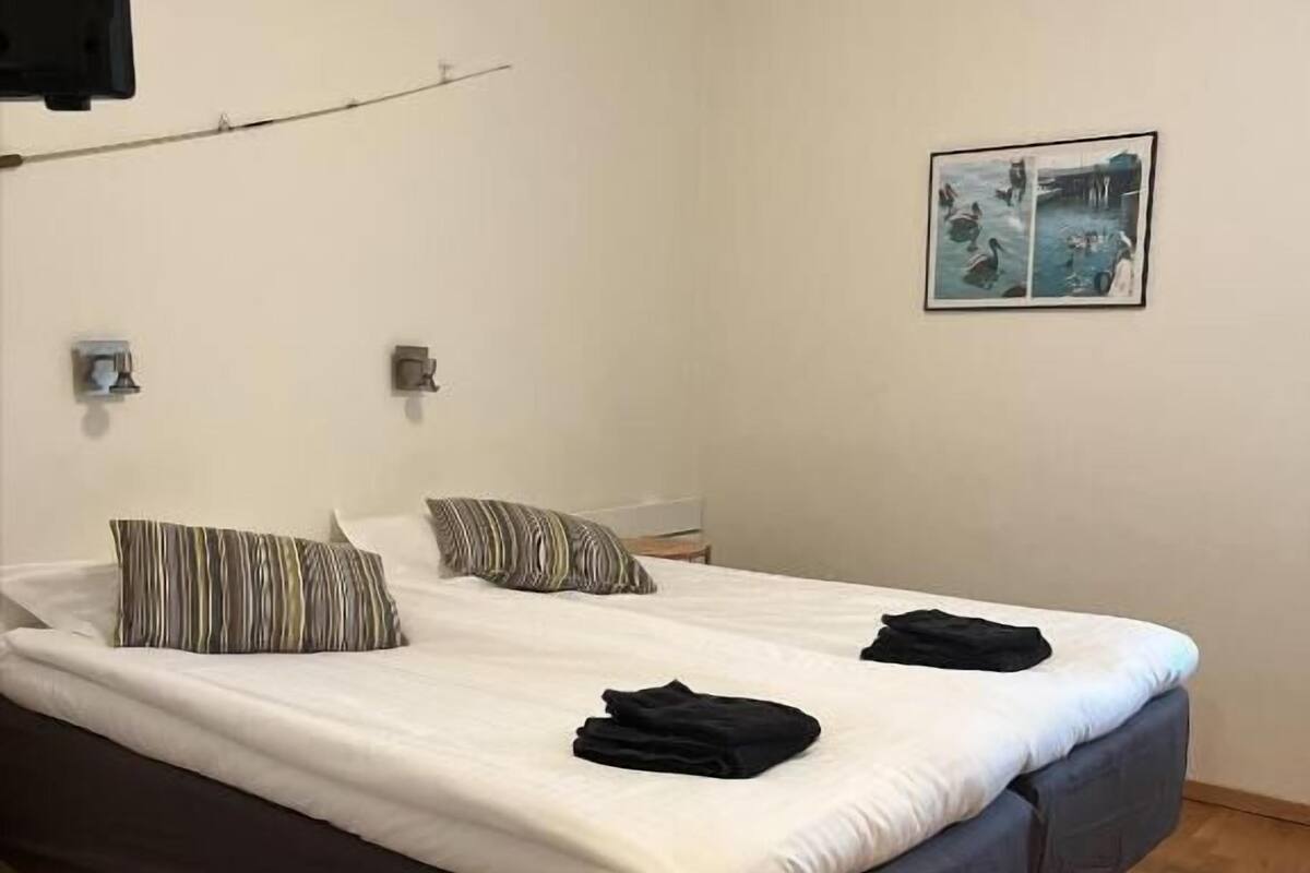 A double bed with a dark headboard, two pillows, and a folded black garment on the bed. There is a framed picture on the wall and a small table beside the bed.