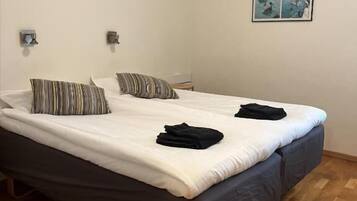 A double bed with a dark headboard, two pillows, and a folded black garment on the bed. There is a framed picture on the wall and a small table beside the bed.