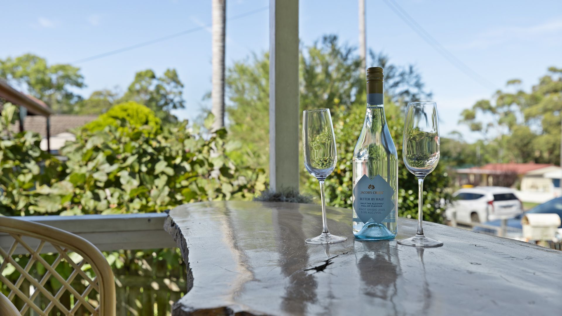 Une table avec une bouteille et deux verres, placée à l’extérieur, avec de la verdure et une voiture en arrière-plan.