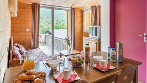 Une table en bois accueillante, dressée pour le petit-déjeuner avec des croissants, du jus de fruits et un saladier rempli de fruits, donnant sur un balcon offrant une vue magnifique.